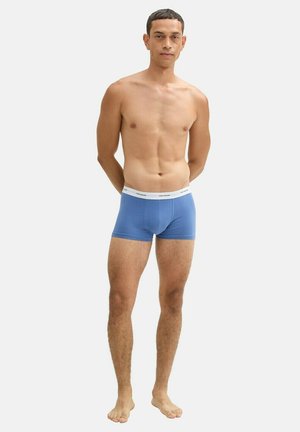 Boxers en coton bleu avec une taille blanche portant un texte noir. Le mannequin se tient pieds nus avec les mains derrière le dos.
