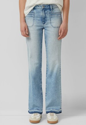 Person trägt hellblaue Bootcut-Jeans mit vorderen aufgesetzten Taschen und weißen Turnschuhen, steht vor einem einfarbigen Hintergrund.