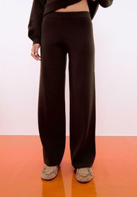 Persona che indossa pantaloni a maglia larghi di colore marrone scuro e scarpe stile mocassino beige, in piedi su un pavimento arancione.