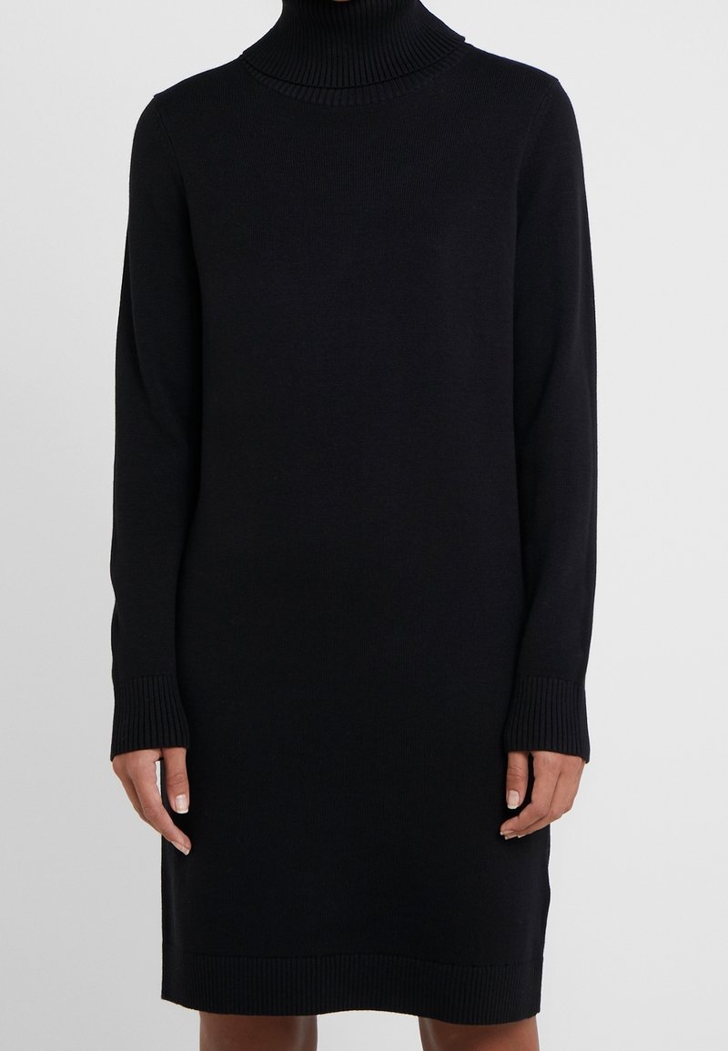 Robe noire à manches longues en tricot côtelé avec col roulé portée par une personne, avec poignets et ourlet côtelés, sur fond clair uni.