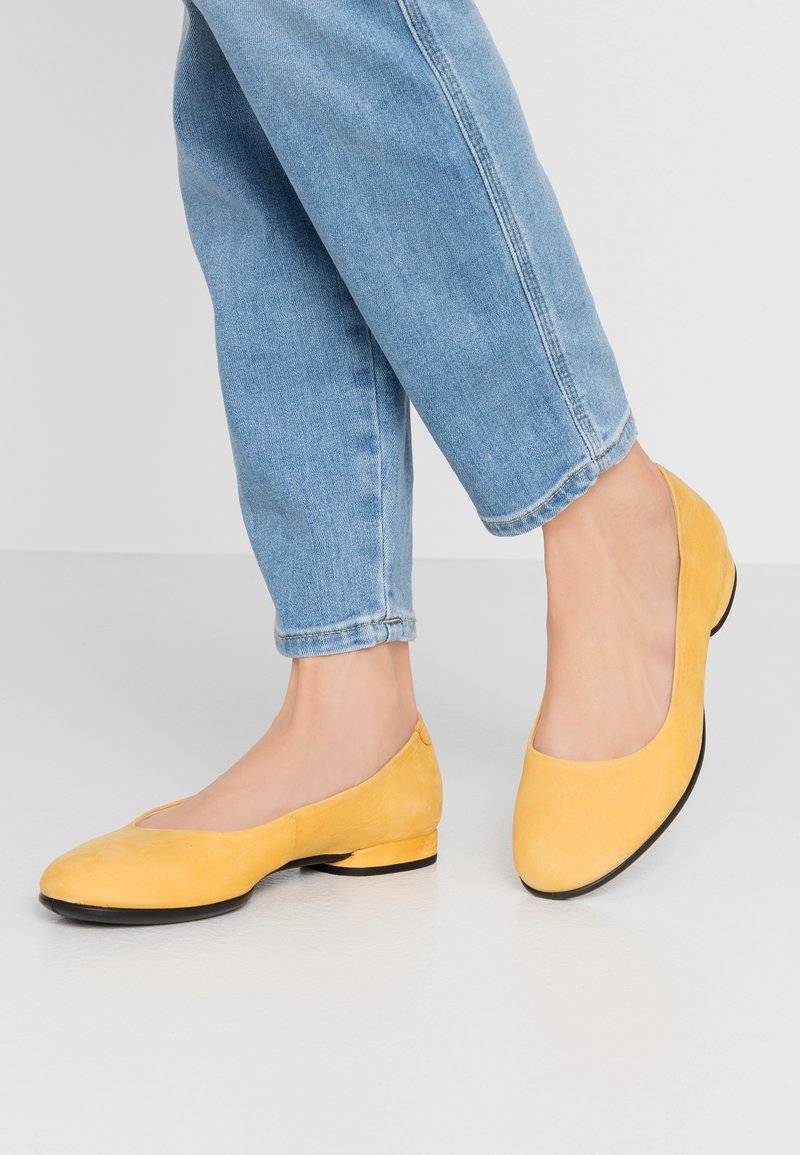 Pieds portant des chaussures basses en daim jaune associées à un jean denim bleu clair raccourci sur un sol gris clair.