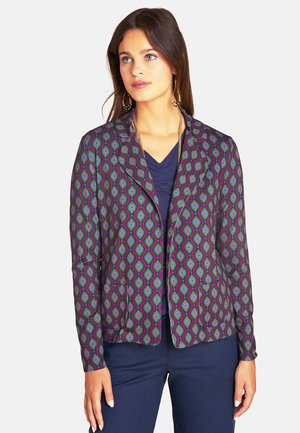Woman with long dark hair wearing gold earrings and a colorful patterned blazer over a dark blue top and matching pants, looking to the side.