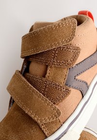 Bruine hoge sneaker met twee verstelbare klittenbandsluitingen, stof- en suède texturen, grijze zijaccent en witte rubberen zool.