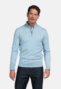 Lichtblauwe gebreide trui met een ritssluiting bij de kraag en een geribde zoom. Draagt over een patroonshirt. Gecombineerd met donkere jeans, die minimale textuur tonen.
