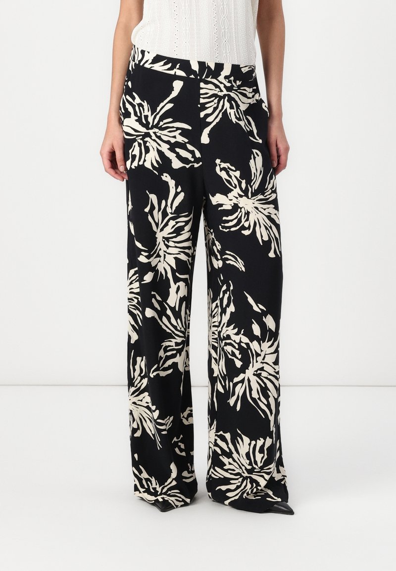 Femme portant un pantalon noir taille haute à jambes larges avec un grand imprimé floral blanc et un haut blanc sans manches.