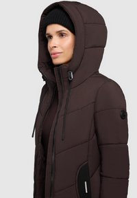Vrouw in een donkerbruine gewatteerde winterjas met capuchon en een zwarte gebreide muts, die naar rechts kijkt tegen een effen lichte achtergrond.