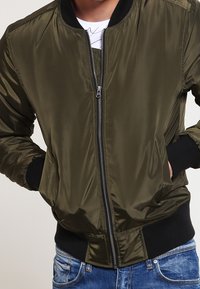 Osoba ve svetle olivově zelené bomber bundě na zip s černými manžetami a pasem, spárována s modrými džínami. Ruce v kapsách bundy.