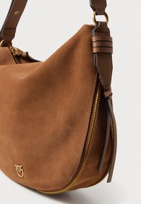 Brūna zamšādas rokassoma ar noapaļotu formu, zelta rāvējslēdzēja detaļām un ādas siksnas akcentiem. Priekšpusē ir gravēts logo.