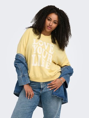 Kvinde med krøllet hår iført en gul sweatshirt med teksten "LOVE YOUR LIFE", blå jeans og en delvist båret denimjakke.