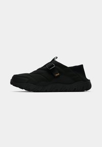 Černé slip-on tenisky s prošívaným svrchním materiálem, nastavitelným popruhem s přezkou a silnou texturovanou podrážkou na čistém bílém pozadí.
