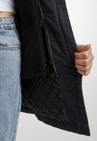 Črna jakna z quiltano notranjostjo in mehkim, teksturiranim zunanjim slojem. Ima zadrgo na strani in stoječ ovratnik.