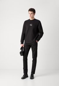 Sweatshirt noir texturé avec logo, associé à un pantalon noir ajusté et des chaussures noires. L'individu tient un sac noir.