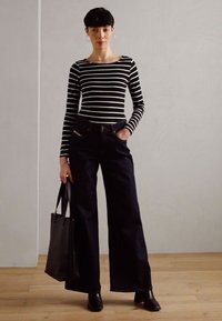Gestreiftes Schwarz-Weiß Langarmhemd, kombiniert mit dunklen weit geschnittenen Jeans. Eine schwarze Schultertasche und spitze Knöchelstiefel runden das Outfit ab.