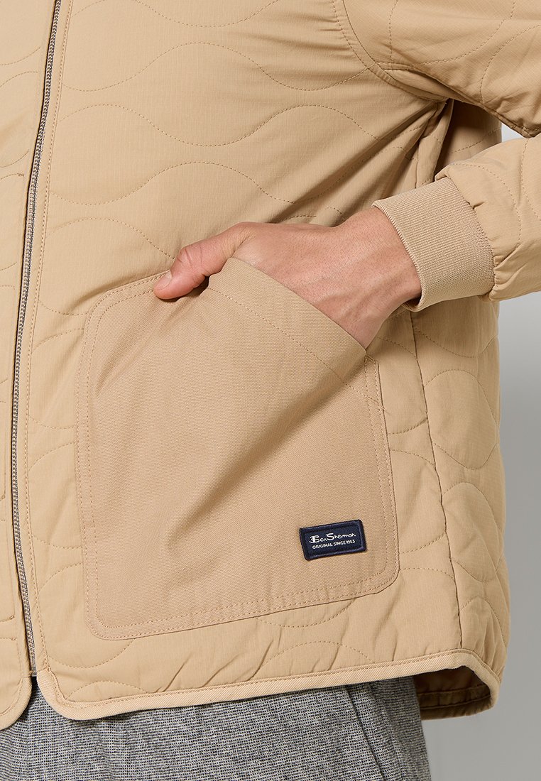 Giacca trapuntata beige con una grande tasca quadrata e un'etichetta ricamata discreta. La texture morbida e i polsini a coste ne aumentano la funzionalità.