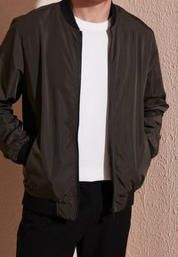 Mies, joka käyttää tummanvihreää pommikakkua valkoisen tekstuuripaidan päällä, kädet takin taskuissa teksturoitua seinää vasten.