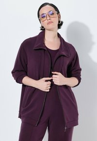 Chaqueta morada con cremallera, hecha de un tejido suave, que presenta un cuello alto y bolsillos laterales, combinada con un conjunto de parte superior e inferior a juego.