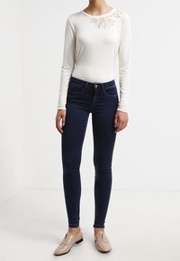 Vit långärmad topp med spetsdetalj vid axeln, parat med mörkblå skinny jeans och beigea slip-on skor.