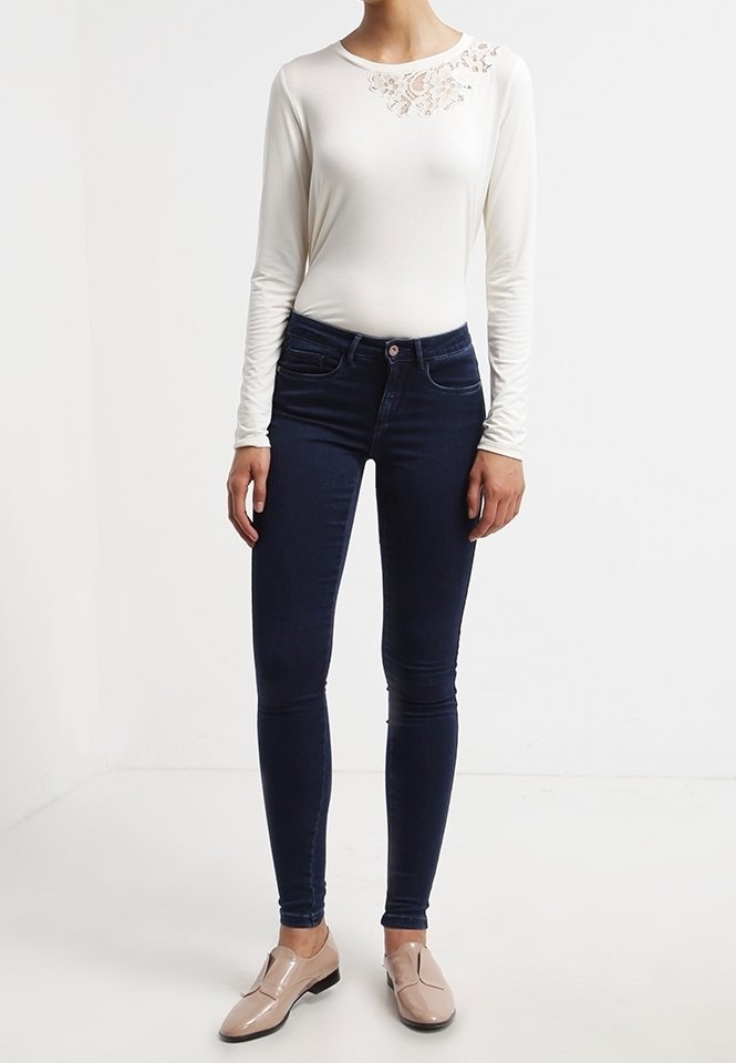 Vit långärmad topp med spetsdetalj vid axeln, parat med mörkblå skinny jeans och beigea slip-on skor.