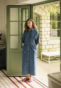 Denimist kleit, keskpikk, nööpidega kinnitusega, küljetaskutega, keered käistega ja lõdva sobivusega. Seljas roosad flip-flop sandaalid.