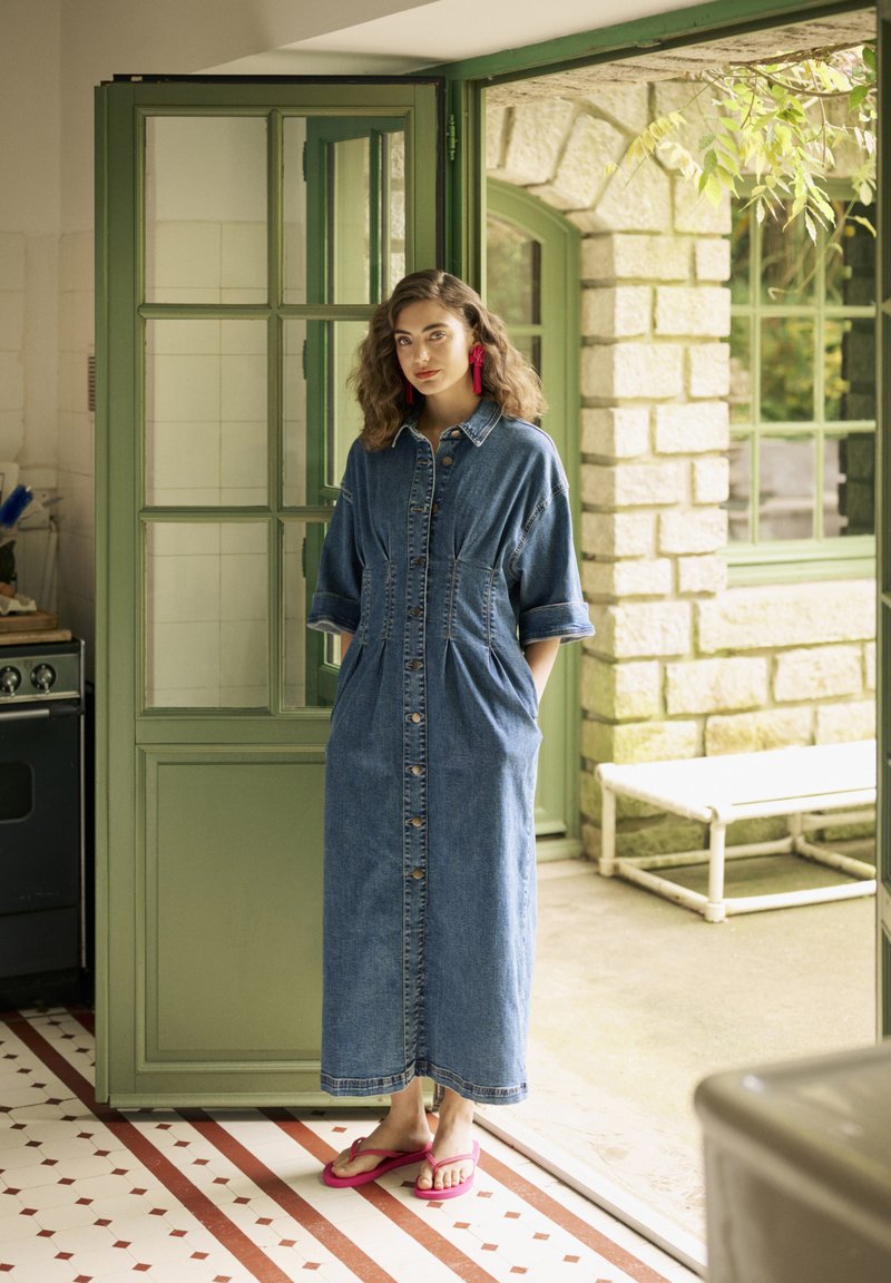 Denimist kleit, keskpikk, nööpidega kinnitusega, küljetaskutega, keered käistega ja lõdva sobivusega. Seljas roosad flip-flop sandaalid.