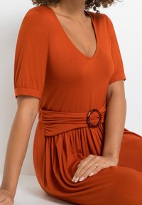 Rdzawopomarańczowa sukienka z dekoltem w serek, krótkimi bufiastymi rękawami, zebranym w talii pasem z okrągłą, żółwiową klamrą i zwiewnym, teksturowanym materiałem.