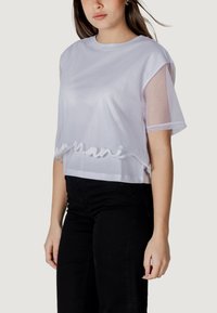 Camiseta corta blanca con un cuerpo liso y mangas cortas de malla translúcida. Presenta el nombre de la marca en textura blanca a lo largo del dobladillo.