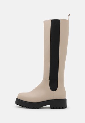 Beiger kniehoher Stiefel mit schwarzem elastischem Seitenteil und klobiger schwarzer Sohle, seitlich auf weißem Hintergrund dargestellt.