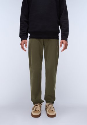 Mann trägt olivgrüne Hose, beige Sneaker mit olivfarbenen Details und ein schwarzes Langarmoberteil, steht vor einem schlichten hellen Hintergrund.