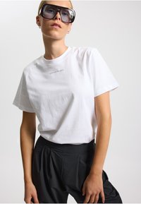 Hvid bomulds-T-shirt med et lille gråt logo, parret med sorte plisserede bukser, der viser en afslappet pasform og blød tekstur.