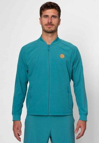 Chaqueta azul verdoso con cremallera y cuello alto, dos bolsillos laterales y un logo circular naranja en el lado izquierdo del pecho. Textura de tela suave.