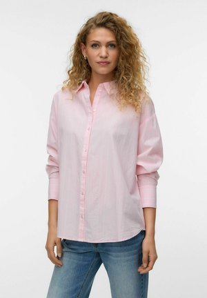 Frau mit lockigem Haar, die ein locker sitzendes hellrosa Hemd mit Knöpfen und blaue Jeans trägt, steht vor einem schlichten weißen Hintergrund.