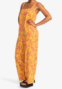 Mono con estampado floral en rosa y amarillo, con piernas anchas, tirantes ajustables y un ajuste relajado; combinado con sandalias de plataforma negras y gruesas.