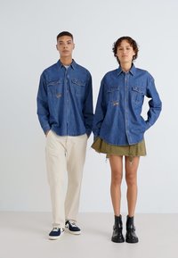 Blå denimskjorter med frontlommer, parret med lysebeige bukser og olivengrøn nederdel. Sorte ankelstøvler og marineblå sneakers fuldender looket.