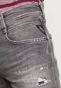 Vaqueros de mezclilla gris con una textura desgastada, costuras visibles y una pequeña etiqueta de marca negra en el bolsillo trasero.