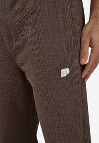 Brune teksturerte bukser med et ribbet design og en hvit brodert "P"-logo på låret. Nærbilde viser stoffvev og form.