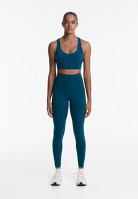 Tengerés színű sportmelltartó és magas derekú leggings rugalmas anyagból, testhezálló kialakítással és sima textúrával. Fehér sportcipők tartoznak hozzá.