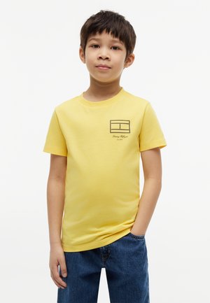 Niño de pie con una mano en el bolsillo, lleva una camisa amarilla de manga corta con un pequeño logo y vaqueros azules sobre fondo blanco.
