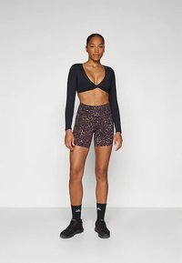 Czarny krótki top z długim rękawem i twistowym wzorem, w połączeniu z brązowymi spodenki rowerowymi w panterkę. Model nosi czarne trampki i skarpety.