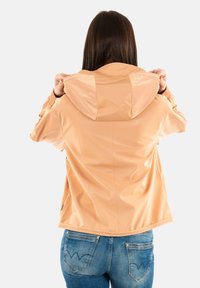 Veste de pluie couleur pêche avec capuche, texture lisse et brillante, poignets élastiques et ourlet légèrement ajusté ; portée avec un jean en denim bleu.