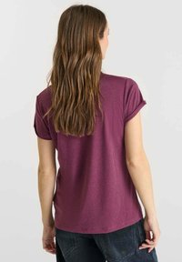 Vrouw met lang bruin haar, gekleed in een paarse T-shirt met korte mouwen en donkere spijkerbroek, staande tegenover een lichtgrijze muur.