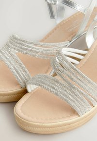 Sølvdekorerede sandaler med flere glitrende remme, blød beige fodseng og en tekstureret gummisål. Har et glat og strømlinet design.