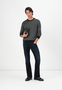 Grauer Strickpullover mit schwarzen Akzenten, kombiniert mit dunkelblauen Schlaghosen. Das Model hält einen schwarzen Gegenstand und steht vor einem schlichten Hintergrund.