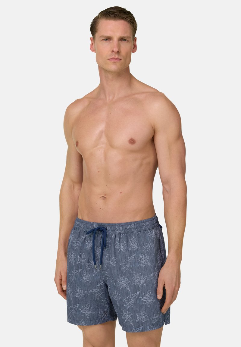 Homme en forme portant un short de bain bleu à motif floral avec un cordon de serrage bleu marine, debout devant un fond clair uni, face à l'avant.