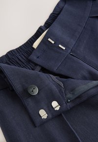 Navyblaue Hose aus strukturiertem Stoff mit elastischem Bund, Knopfverschluss, silberner Hardware und einem Reißverschlusstaschen-Detail.