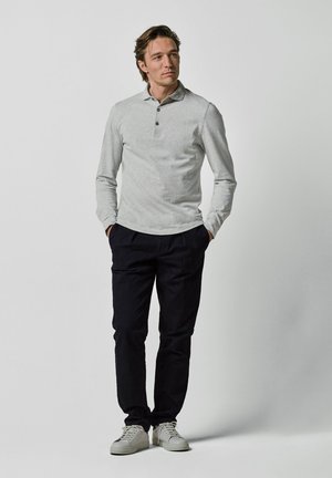 Mann trägt ein hellgraues langärmeliges Polohemd, schwarze Hose und weiße Sneakers, steht mit den Händen in den Taschen vor einem schlichten Hintergrund.