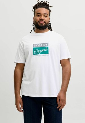 Szakállas, raszta hajú férfi fehér "Jack & Jones Originals" pólóban és sötét nadrágban, egyszínű világos háttér előtt áll.