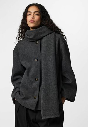 Femme aux longs cheveux bouclés portant un manteau gris foncé boutonné avec une écharpe attachée et un pantalon noir sur un fond uni.