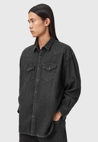 Czarna koszula dżinsowa z kołnierzykiem, z zapięciem na guziki, dwoma kieszeniami na piersi i długimi rękawami. Tkanina ma lekko teksturowane wykończenie.