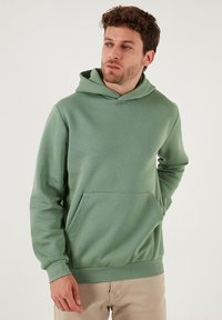 Vihreä huppari pehmeästä kankaasta, jossa etutasku, resorit hihansuissa ja nyörillinen huppu. Sileä pinta, tavallinen istuvuus.