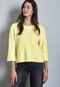 Frau mit langem dunklem Haar, die einen hellgelben Strickpullover mit drei Viertelärmeln und schwarze Jeans trägt, steht vor einer grauen Wand.